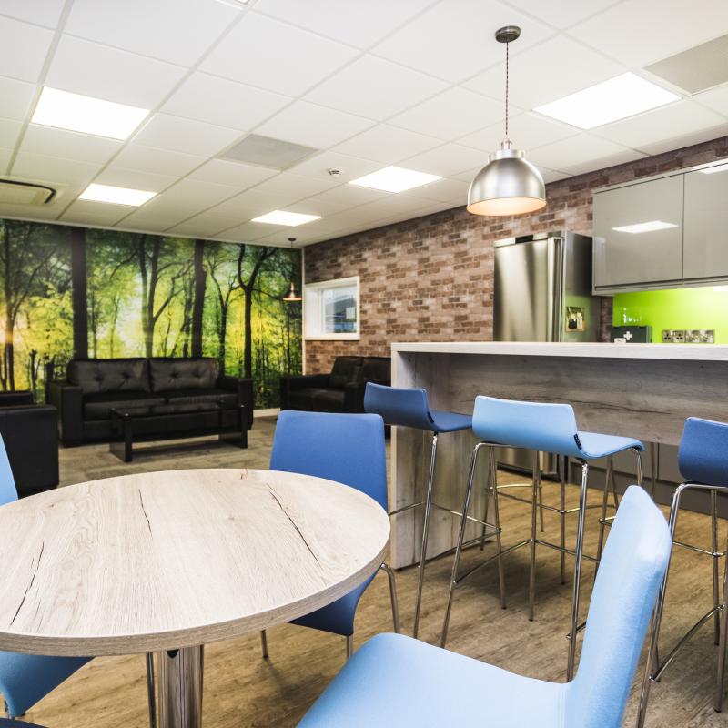 image of ground floor breakout / tea-point and lounge area with light oak coloured round table and high bench, two different blue chairs and stools in front of a straight section of gloss grey kitchen units. The units have a lime green splash-back. The lounge area has three  black leather sofas around black glass coffee table. Surrounding the sofas is a large forest scene image across the walls. There is feature pendant lighting above the high bench and in the lounge area corners.