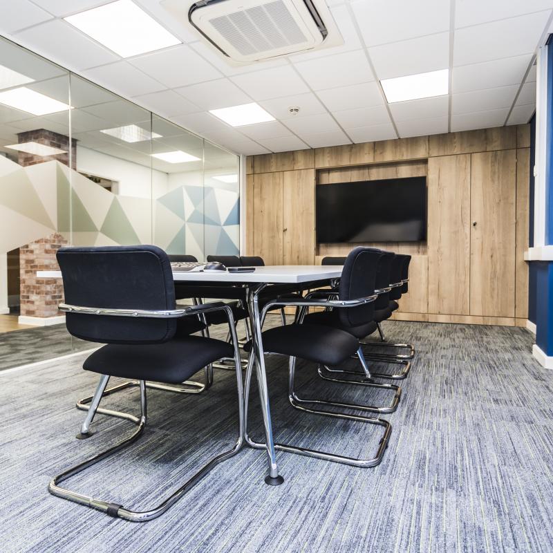 Image of ground floor meeting room showing white table, black and chrome chairs. Blue wall to right, end wall is a media panel in oak with large TV screen in the middle. Left wall of meeting room is glazed floor to ceiling with green and blue geometric shaped branding across central area.