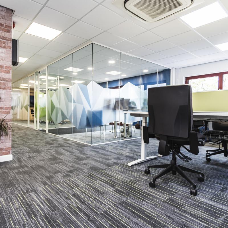 Image of ground floor desks looking towards the entrance foyer and meeting room. Black and grey carpet with green lines under desks and black lines for main corridor. Brick column with plant. Branding graphics on glazed walls.