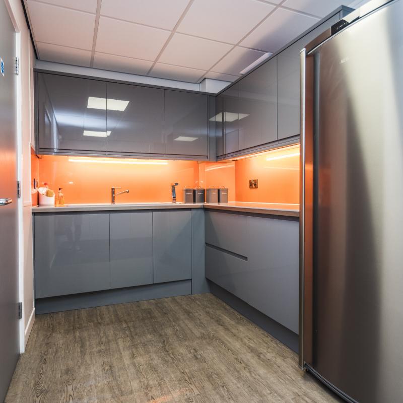 Image of 1st floor tea point showing grey gloss units with high gloss orange splash back. Pelmet lights on under wall units creating reflections on orange splash back, Wooden plank effect flooring and stainless steel fridge.