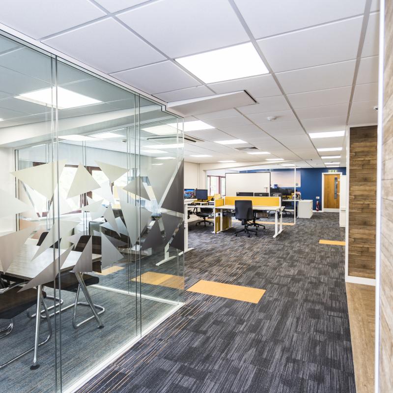 Image of bright new 1st floor open plan office with MD's glazed office on left showing new black and orange branding. Carpet is black and grey with small orange lines and inset orange tiles. Desks are white with orange screens and grey chairs.