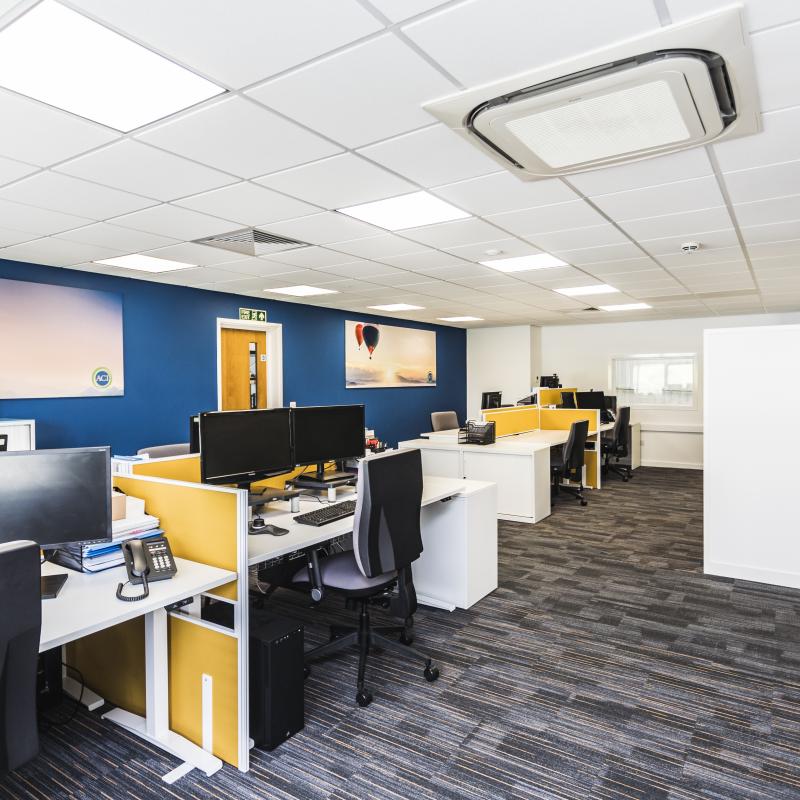 Image of 1st floor desk area looking towards mezzanine exit. View shows two banks of white desks with orange screens and grey chairs. Also white side panel of printer area. Rear wall is dark blue with two large horizontal pictures showing balloon scenes.